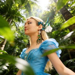 May include: A person in a blue gown with a feathered hairpiece and peacock feather earrings stands in a verdant forest. The gown has a sweetheart neckline and sheer sleeves. The hairpiece and earrings feature blue, green, and white feathers.