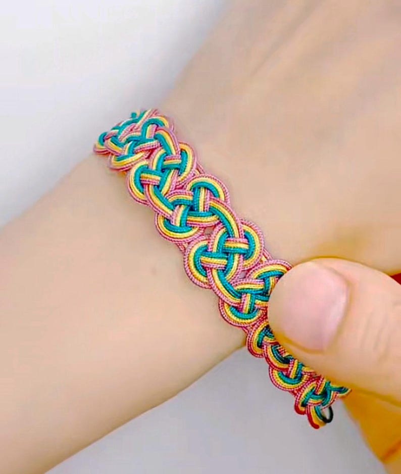 May include: A braided bracelet with pink, yellow, and teal threads woven together in a complex pattern.