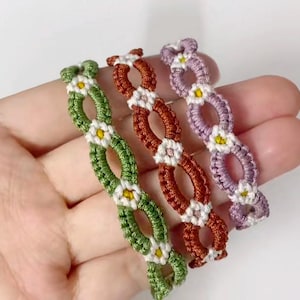 Pode incluir: Três pulseiras de amizade com um design de margarida. As pulseiras são verdes, laranja e roxas.