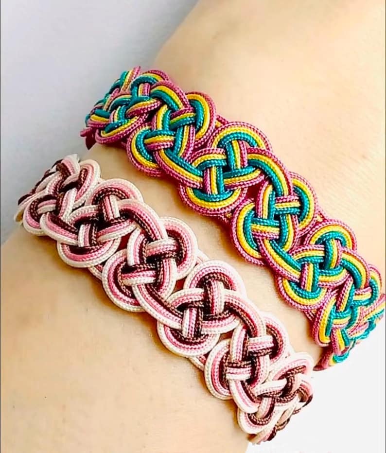 May include: Two braided bracelets, one with pink, white, and brown cords, and the other with pink, yellow, and teal cords.