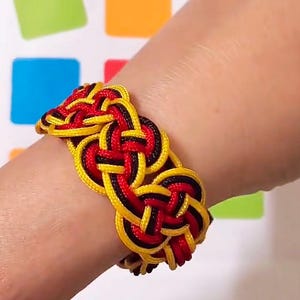 May include: A black, red, and yellow braided bracelet with a knot pattern. The bracelet is worn on a person's wrist.
