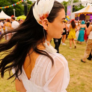 May include: A white feather headpiece with red and brown accents, worn on the side of the head. A matching feather earring hangs from the ear. The person is wearing a white dress at an outdoor event.