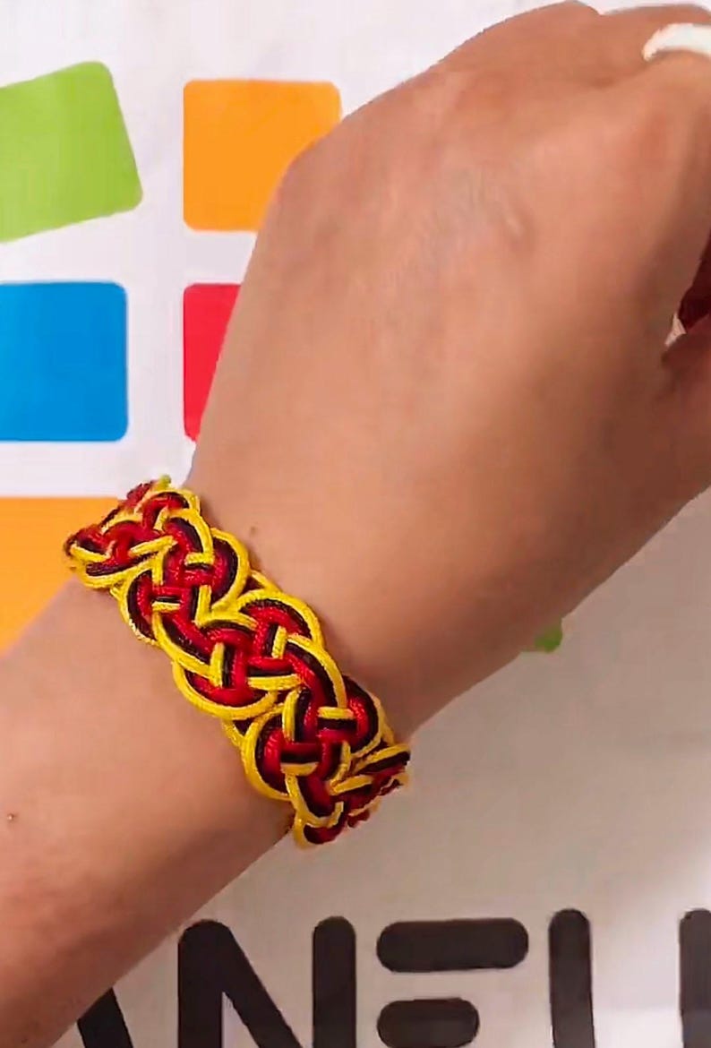 May include: A black, red, and yellow braided bracelet. The bracelet is made of woven string and is worn on a person's wrist.