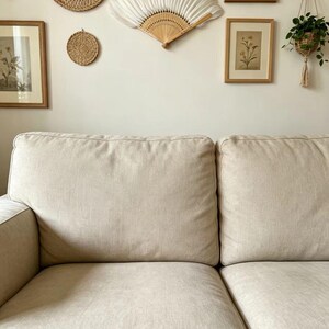 May include: A beige sofa with two large cushions dominates the foreground. The wall behind the sofa is adorned with a round woven mirror, a small woven coaster, a white fan, framed botanical prints, and a hanging plant.