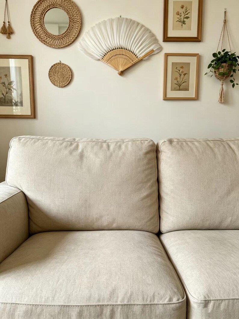 May include: A beige sofa with two large cushions dominates the foreground. The wall behind the sofa is adorned with a round woven mirror, a small woven coaster, a white fan, framed botanical prints, and a hanging plant.