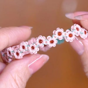 May include: A pink and green friendship bracelet with small pink flowers and orange centers.