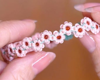 May include: A pink and green friendship bracelet with small pink flowers and orange centres.