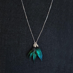 Könnte beinhalten: Eine silberne Halskette mit feiner Kette. Der Anhänger zeigt eine Ansammlung von geschichteten, irisierenden grün-blauen blattförmigen Elementen. Die Halskette wird vor einem dunklen Hintergrund präsentiert.