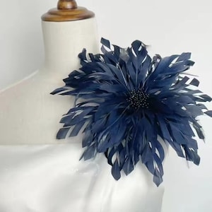 May include: A navy blue feather flower accessory, possibly a brooch or hair clip, is displayed on a mannequin. The flower has a central beaded detail and layered feathers, creating a full, textured appearance. The accessory is likely for formal wear.