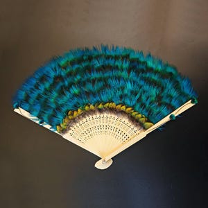 May include: A handheld folding fan featuring vibrant teal and blue feathers layered across the top. The base consists of light-colored, intricately carved wooden ribs that radiate from a central pivot point, creating a delicate, open structure.