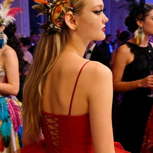 May include: A woman in a red formal dress with a corset back and spaghetti straps. She wears a feathered hairpiece with orange, brown, and black feathers. Other women in the background also wear feathered accessories. The event appears to be a formal ball.