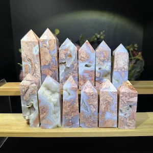 May include: A collection of 12 pink and white agate crystal points, arranged in a row on a wooden shelf. The points are all different sizes and shapes, and they have a natural, rough texture.