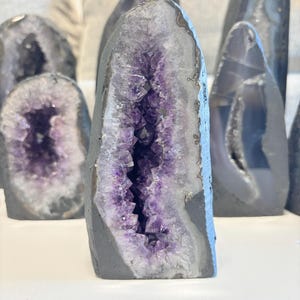 May include: Several amethyst geodes are displayed, varying in size and form. Each geode has a grey outer layer, revealing a bright purple crystal interior. The crystals glisten, offering a striking visual. These natural formations are presented against a plain backdrop.