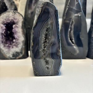May include: A polished, dark blue agate geode with white and grey banding. The geode has a hollow centre filled with sparkling crystals. Other geodes are visible in the background, showcasing a collection of natural stone specimens.
