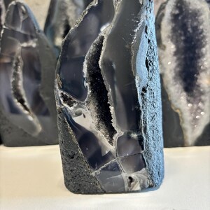 May include: A dark grey agate geode with a rough, textured exterior and a polished, reflective interior. The geode has a jagged, irregular shape, with a cavity lined with sparkling crystals. Other geodes are visible in the background.