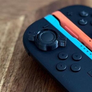 May include: Black game controller with orange and blue accents. The controller has buttons, joysticks, and a central connector. Black cords extend from the bottom. The controller is on a wooden surface.