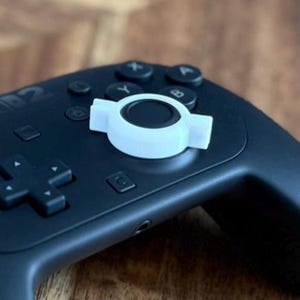 May include: A black video game controller with white accents on the joysticks and buttons. The controller is resting on a wooden surface with a natural grain pattern. The controller has a sleek, ergonomic design.
