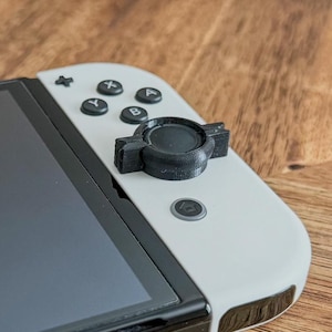 May include: A black 3D-printed accessory attached to a white game controller. The accessory is circular with a notched design. The controller has black buttons and a screen, resting on a wooden surface.