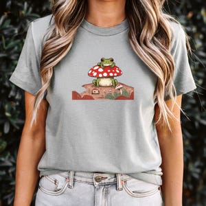 May include: A light gray short-sleeve t-shirt featuring a graphic print of a green frog sitting on a large red mushroom with white spots. The illustration includes smaller mushrooms and earthy ground details in shades of brown and green.