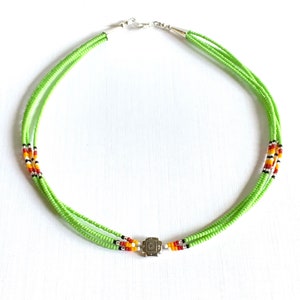 Op de afbeelding: Een meerstrengs ketting gemaakt van groene zaadkraaltjes met een zilveren sluiting en een zilveren accentkraal. De ketting heeft een klein gedeelte met rode, oranje en zwarte kralen.