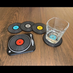 Könnte beinhalten: Satz von sechs schwarzen Vinyl-Untersetzern in Form von Schallplatten mit einem Plattenspieler-Design. Ein Untersetzer zeigt eine rote Schallplatte, während die anderen blaue, gelbe und graue Schallplatten haben. Die Untersetzer befinden sich auf einer Holzoberfläche.