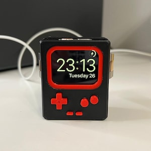 May include: A black and red 3D printed case for an Apple Watch with a digital clock face displaying "23:13 Tuesday 26" and a retro-style game controller design with red buttons.