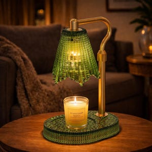 May include: A green glass candle warmer with a brass-colored metal frame. The warmer has a decorative green glass shade and base. A lit candle sits on the base. The candle warmer is on a wooden table.