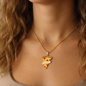 May include: A gold-colored necklace with a pizza slice pendant. The pendant is yellow with red pepperoni and a brown crust. A banner on the pendant reads "TRUE LOVE" in white letters. The necklace is worn around the neck.