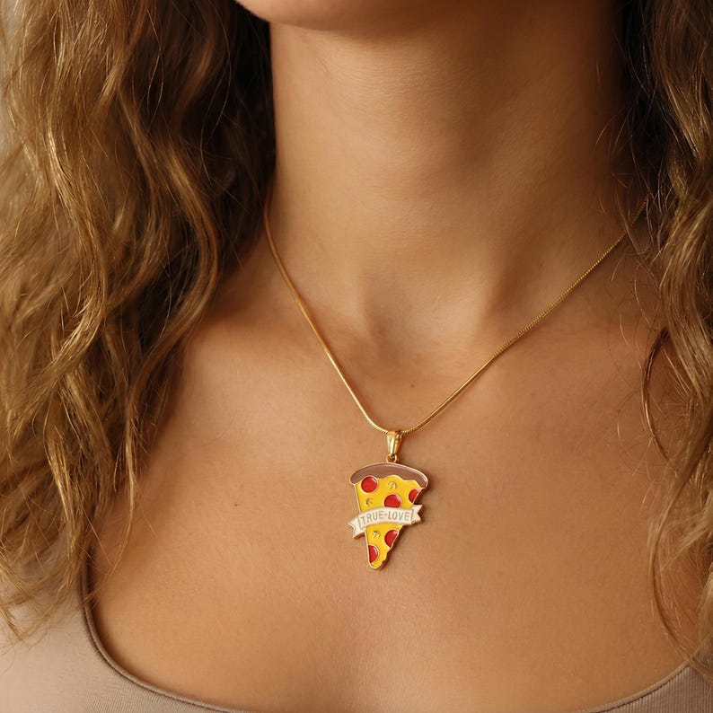 May include: A gold-colored necklace with a pizza slice pendant. The pendant is yellow with red pepperoni and a brown crust. A banner on the pendant reads "TRUE LOVE" in white letters. The necklace is worn around the neck.