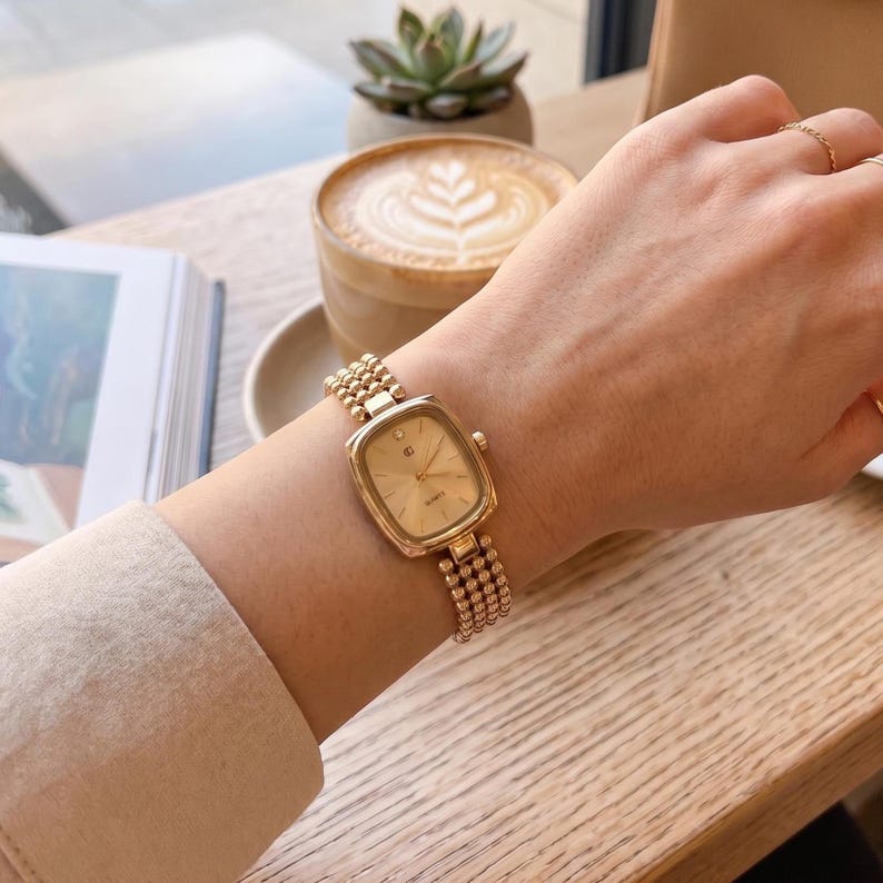 May include: A gold-tone quartz wristwatch featuring a rectangular case with rounded corners and a champagne-colored dial. The watch has a multi-strand beaded bracelet made of small gold-tone spheres, creating a textured, flexible band design.