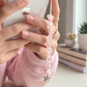May include: A clear phone case with a white cord and a pink gemstone phone charm. The charm features a string of pink and clear stones, and a silver star charm at the end. The phone is held in a person's hands.