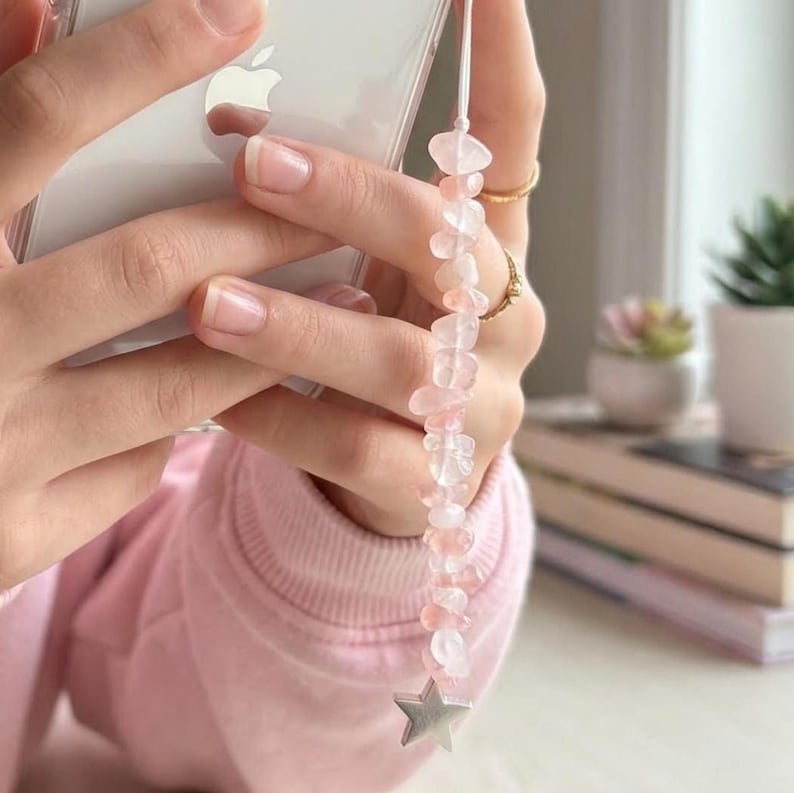 May include: A clear phone case with a white cord and a pink gemstone phone charm. The charm features a string of pink and clear stones, and a silver star charm at the end. The phone is held in a person's hands.