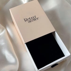 May include: A partially open rectangular box with a light pink lid and white sides. The box interior is lined with black velvet. The text "Retro atelier" is printed on the lid in black. The box is on a white satin surface.