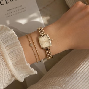 May include: A gold-toned wristwatch with a rectangular face and a beaded metal strap, accompanied by two thin gold-toned bracelets. One bracelet is a simple wire, and the other is a delicate chain featuring a small, circular, stone-encrusted charm.