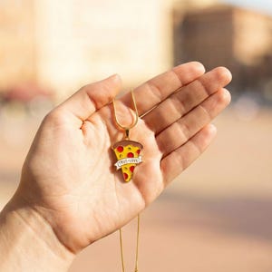 May include: A gold-colored necklace featuring a pizza slice pendant. The pendant has a yellow crust, red pepperoni, and a banner that reads "TRUE LOVE". The necklace is held in a hand.