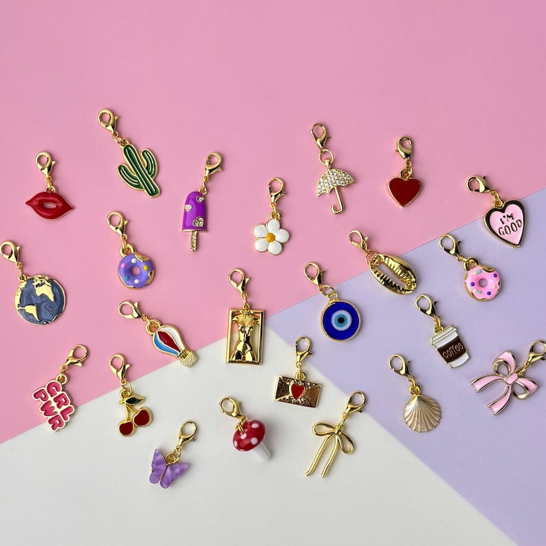 Puede incluir: Una colecci&oacute;n de colgantes dorados con varios dise&ntilde;os, incluyendo un cactus, labios, una rosquilla, una flor, un coraz&oacute;n y una taza de caf&eacute;. Los colgantes se muestran sobre un fondo rosa y morado.