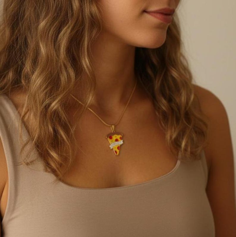 May include: A gold-colored necklace with a pizza slice pendant. The pendant features a yellow base with red and white accents, and a banner with text. The necklace is worn on a person.