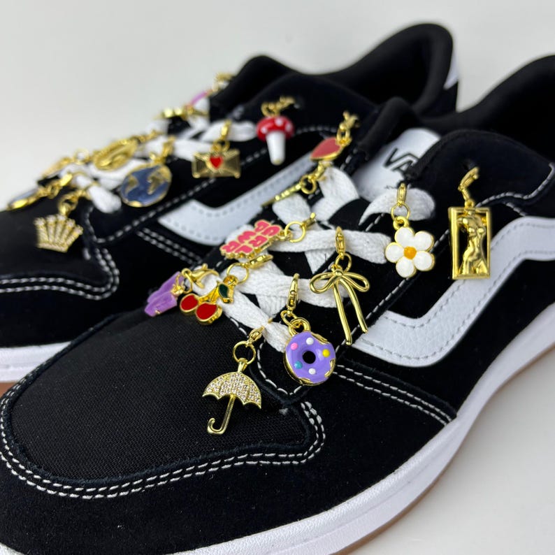 Puede incluir: Zapatillas negras y blancas con cordones blancos adornadas con varios dijes dorados. Los dijes incluyen una corona, un globo terr&aacute;queo, una seta, una cereza, un lazo, una rosquilla y un paraguas. Los zapatos tienen una suela blanca.