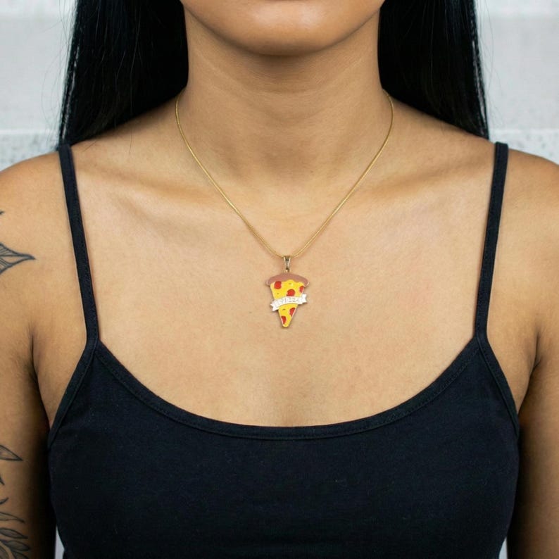 May include: A gold-colored necklace with a pizza slice pendant. The pendant features a slice of pizza with red, yellow, and brown colors. A banner with the word "Pizza" is on the pendant. The necklace is worn on a person wearing a black tank top.