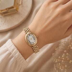 May include: A gold-toned wristwatch featuring an oval-shaped case and a white dial with black Roman numeral hour markers. The watch has a matching gold-toned metal link bracelet and a small crown on the side for time adjustment.