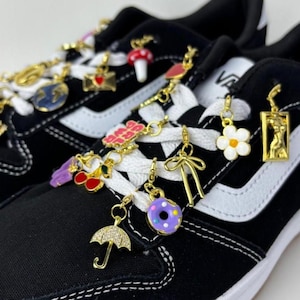 Puede incluir: Zapatillas negras y blancas adornadas con abalorios dorados. Los abalorios incluyen una seta, un coraz&oacute;n, una flor, un lazo y un donut. Las zapatillas tienen cordones blancos y una raya blanca en el lateral.