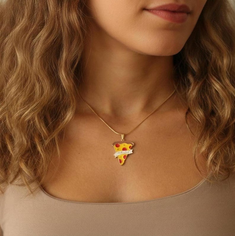 May include: A gold-colored necklace with a pizza slice pendant. The pendant features a yellow crust, red pepperoni, and a banner that reads "TRUE LOVE". The necklace is worn on a person.