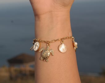 May include: A gold-toned chain charm bracelet featuring multiple dangling pendants, including a sea turtle, a textured seashell, and an oval charm with a white inlay and a cross design, secured with a simple clasp around the wrist.