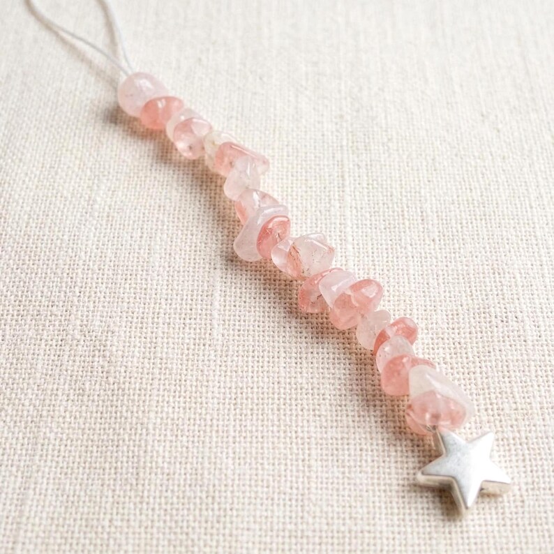 May include: A delicate, handmade gemstone pendant featuring a string of pink and clear quartz chips. The pendant is accented with a silver-colored star charm at the bottom and a white string for hanging. The quartz chips are irregular in shape and size.