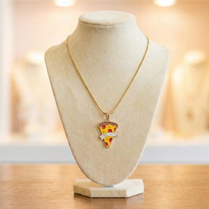 May include: A gold-colored necklace with a pizza slice pendant. The pendant features a slice of pizza with red sauce, yellow cheese, and a crust. A banner on the pendant reads "TRUE LOVE". The necklace is displayed on a mannequin.