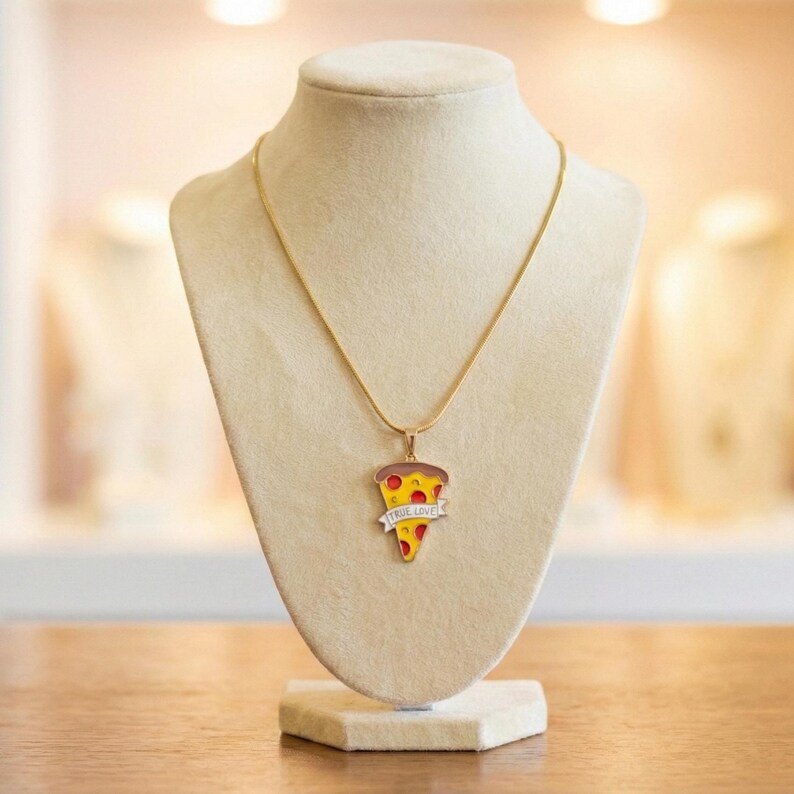 May include: A gold-colored necklace with a pizza slice pendant. The pendant features a slice of pizza with red sauce, yellow cheese, and a crust. A banner on the pendant reads "TRUE LOVE". The necklace is displayed on a mannequin.