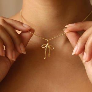 Könnte beinhalten: Eine goldfarbene Halskette mit einem anhänger in Schleifenform. Der Anhänger hat ein minimalistisches Design mit zwei gebogenen Linien, die die Schleifenschleifen bilden, und zwei geraden Linien, die nach unten hängen. Die Halskette wird von einer Person gehalten.