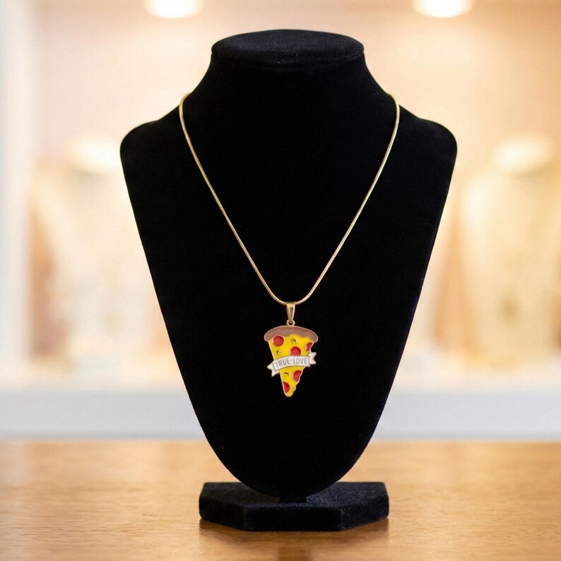 May include: A gold-colored necklace with a pizza slice pendant. The pendant features a slice of pizza with red, yellow, and brown colors, and the words "TRUE LOVE" on a banner. The necklace is displayed on a black jewelry stand.