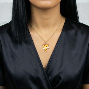 May include: A gold-colored necklace with a pizza slice pendant. The pendant features a slice of pizza with red, yellow, and brown details, and a banner with text. The necklace is worn against a black top.