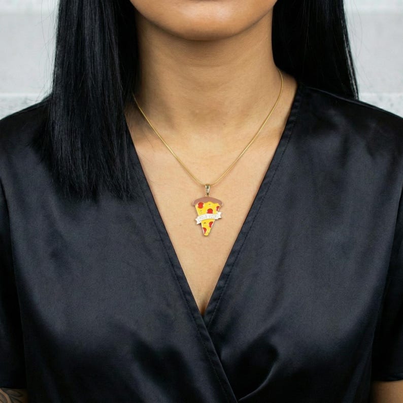 May include: A gold-colored necklace with a pizza slice pendant. The pendant features a slice of pizza with red, yellow, and brown details, and a banner with text. The necklace is worn against a black top.